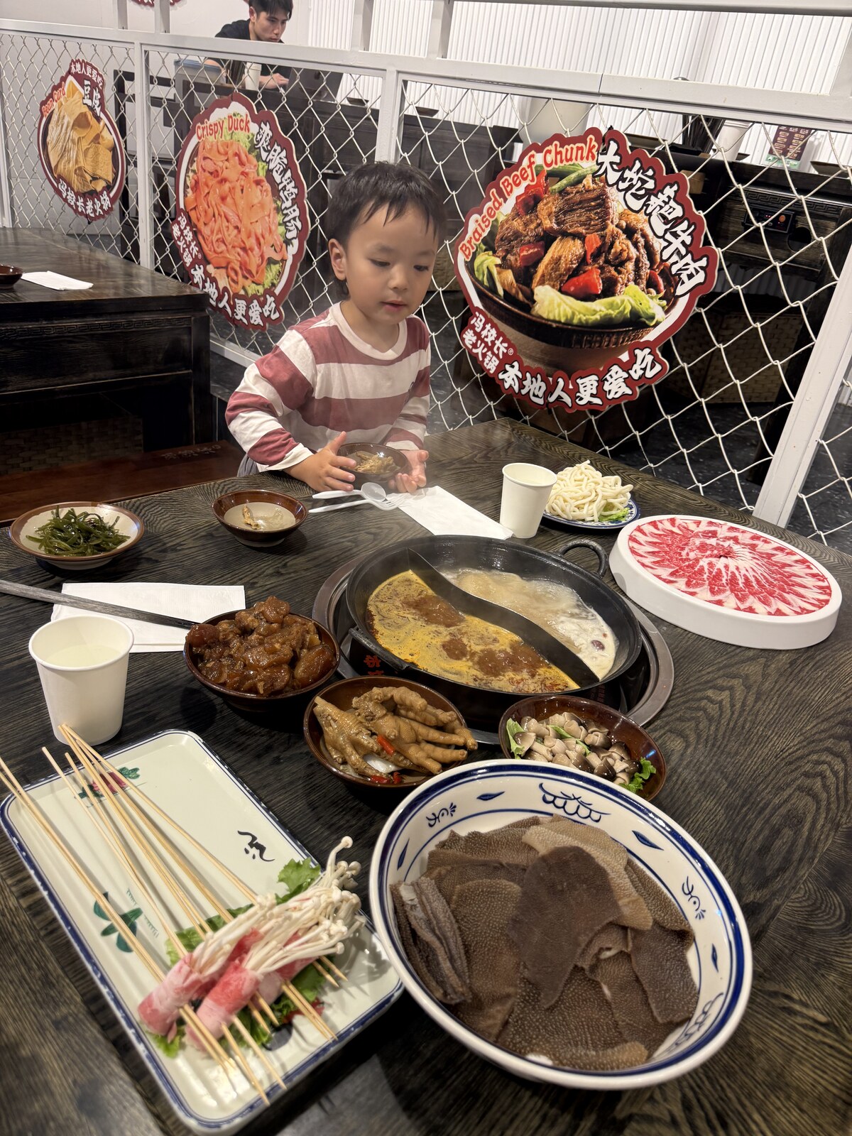 Jichang sitting at a hot pot table with sliced meat, skewers, and soup pots